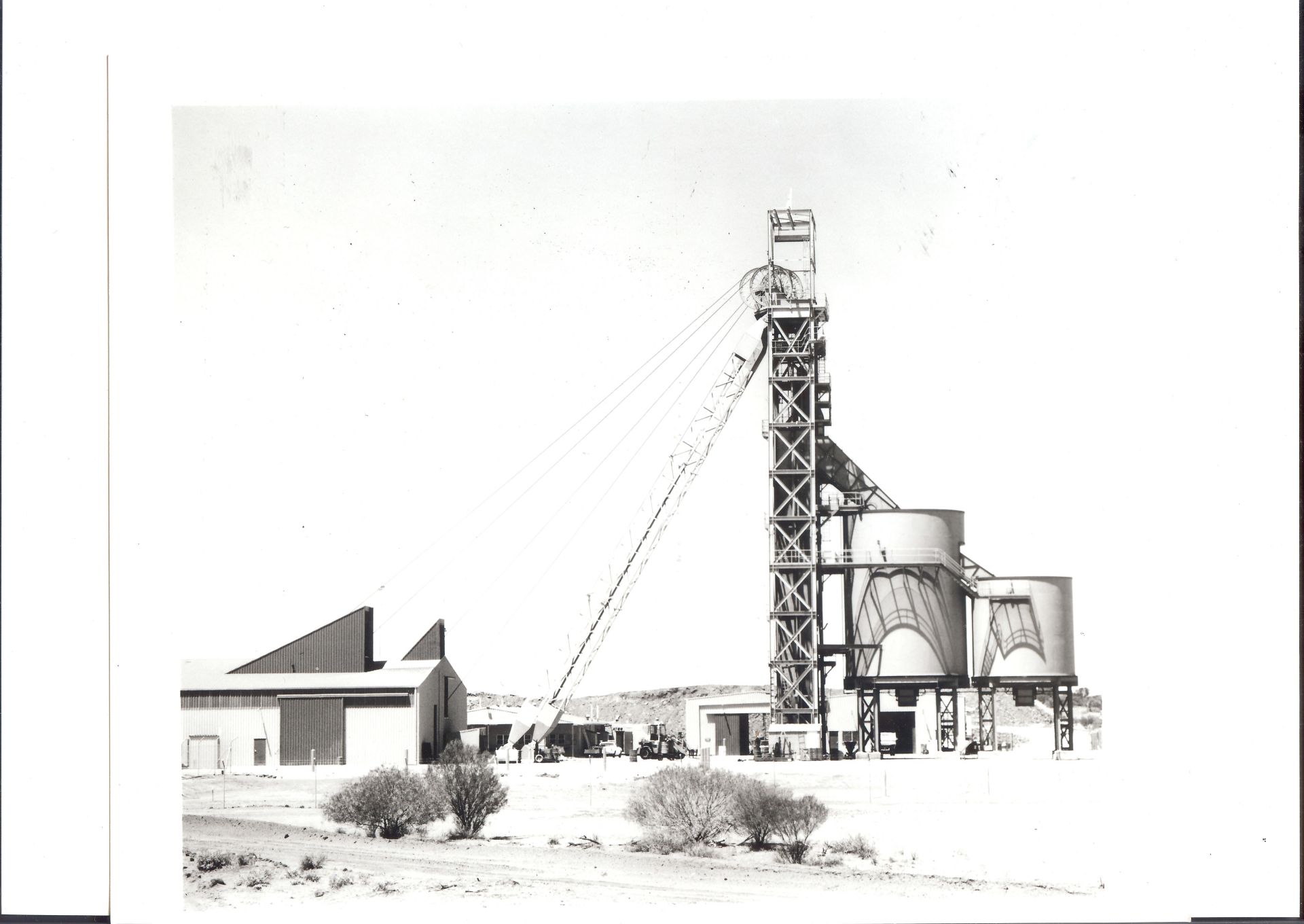 Black-and-white photograph of the Whenan Shaft at the Olympic Dam mine site, showing the towering mining structure, adjacent silos, and nearby buildings, surrounded by a sparse outback landscape.