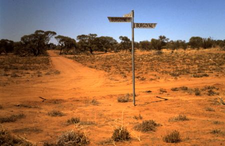 A historical photo showing the original location of Richardson Place, now the main street in Roxby Downs, viewed from what is currently the hospital site. The image features a barren, red desert landscape with sparse vegetation, a dirt path, and a street sign reading 'Burgoyne St.' against a clear blue sky