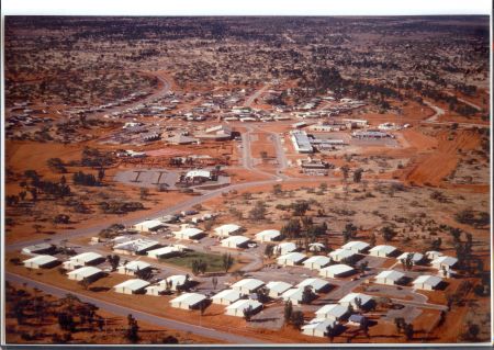 An aerial photograph of Roxby Downs circa 1987-1988, showcasing the town in its early development stages. The image highlights rows of small white-roofed houses, sparse vegetation, and red desert terrain. Key landmarks such as the hospital, auditorium, and houses on Stuart Road are notably absent, reflecting the early days of the town's growth.